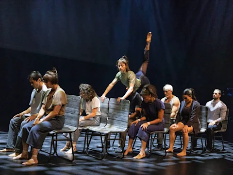 A group of people sit in two rows of chairs on stage, with one person climbing over the chairs and raising a leg into the air. Stage lighting highlights the performers.