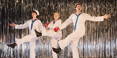 Three performers in white sailor costumes dance in front of a metallic fringe curtain, each lifting one leg in a synchronized, energetic pose.