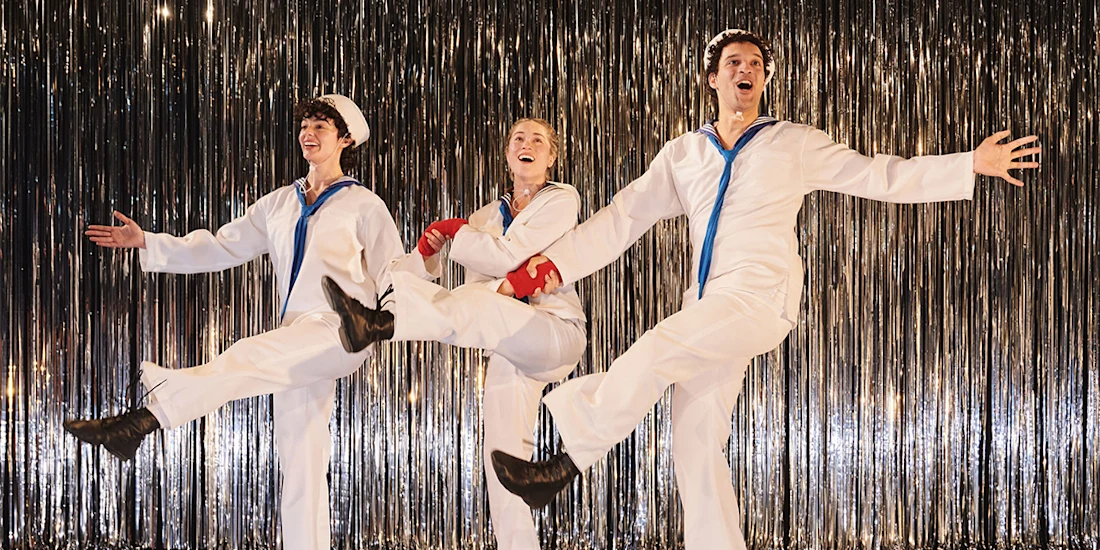 Three performers in white sailor costumes dance in front of a metallic fringe curtain, each lifting one leg in a synchronized, energetic pose.