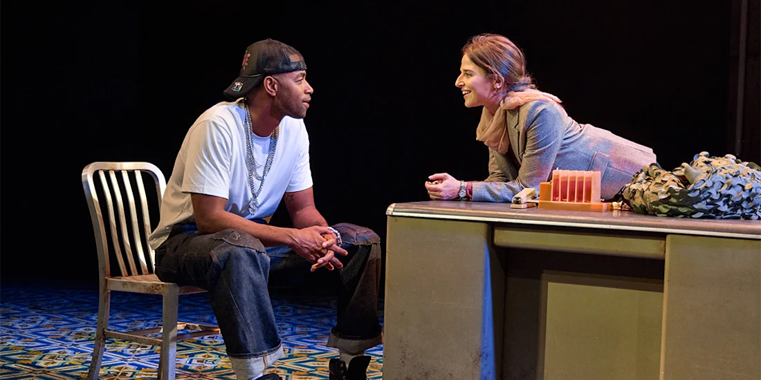 A man in a white t-shirt and cap sits facing a woman who is leaning forward over a desk, both engaged in conversation on a stage set.