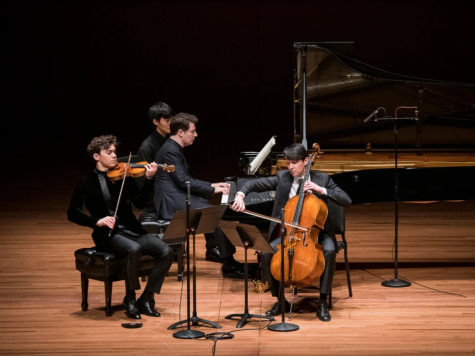 Three musicians perform on stage: one playing a violin, one playing a cello, and one playing a piano. Music stands with sheet music are in front of the string players.