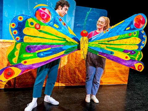 Two people on stage hold a large, colorful butterfly prop with vibrant wings; the background features bright, painted scenery.