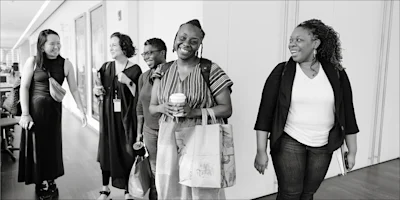 Five women walk together in a hallway, smiling and talking. One holds a coffee cup and tote bag, another carries a notebook. The scene appears to be in an office environment.