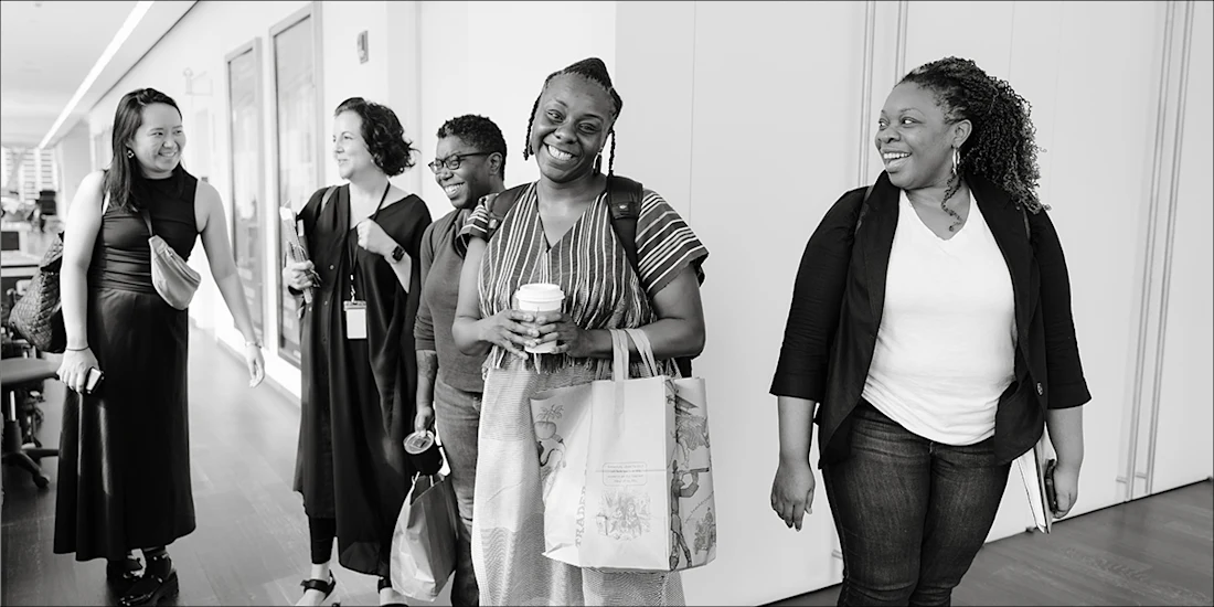 Five women walk together in a hallway, smiling and talking. One holds a coffee cup and tote bag, another carries a notebook. The scene appears to be in an office environment.