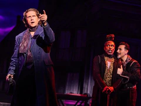 Three actors on stage in an intense scene, with one pointing forward and two others engaged in the background, wearing period costumes. The backdrop is dark with dramatic lighting.