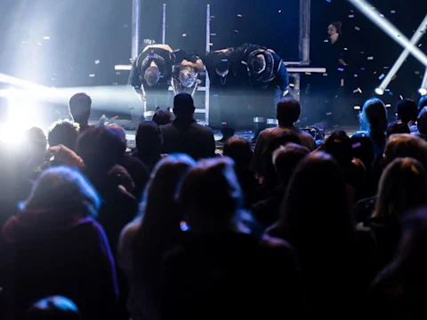 Performers take a bow on a stage in front of an audience, with confetti falling and bright stage lights shining.
