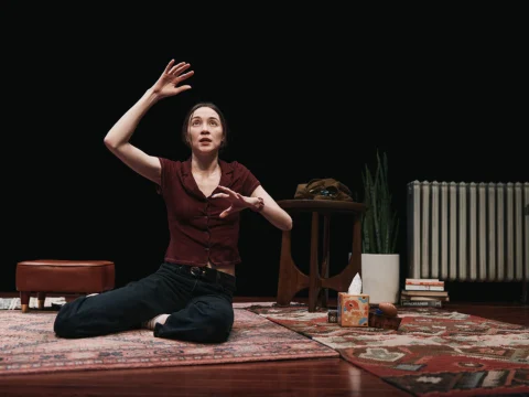 A person sits on a patterned rug, gesturing with one hand raised. A small table with a potted plant, books, tissues, and other items is beside them.