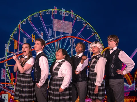 Production shot of Ride the Cyclone in San Francisco, showing ensemble in matching plaid outfits stand in front of a brightly lit Ferris wheel and roller coaster at night, striking playful poses.