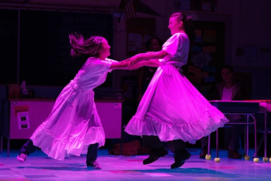 Two people in white dresses hold hands and spin each other in a classroom, both appearing joyful under purple stage lighting.
