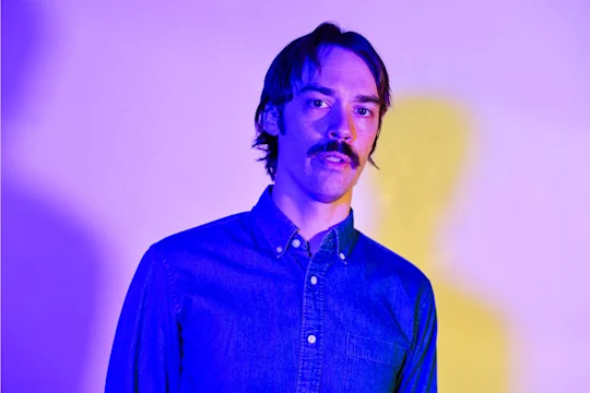 A man with a mustache wearing a button-up denim shirt stands in front of a purple and yellow gradient background.