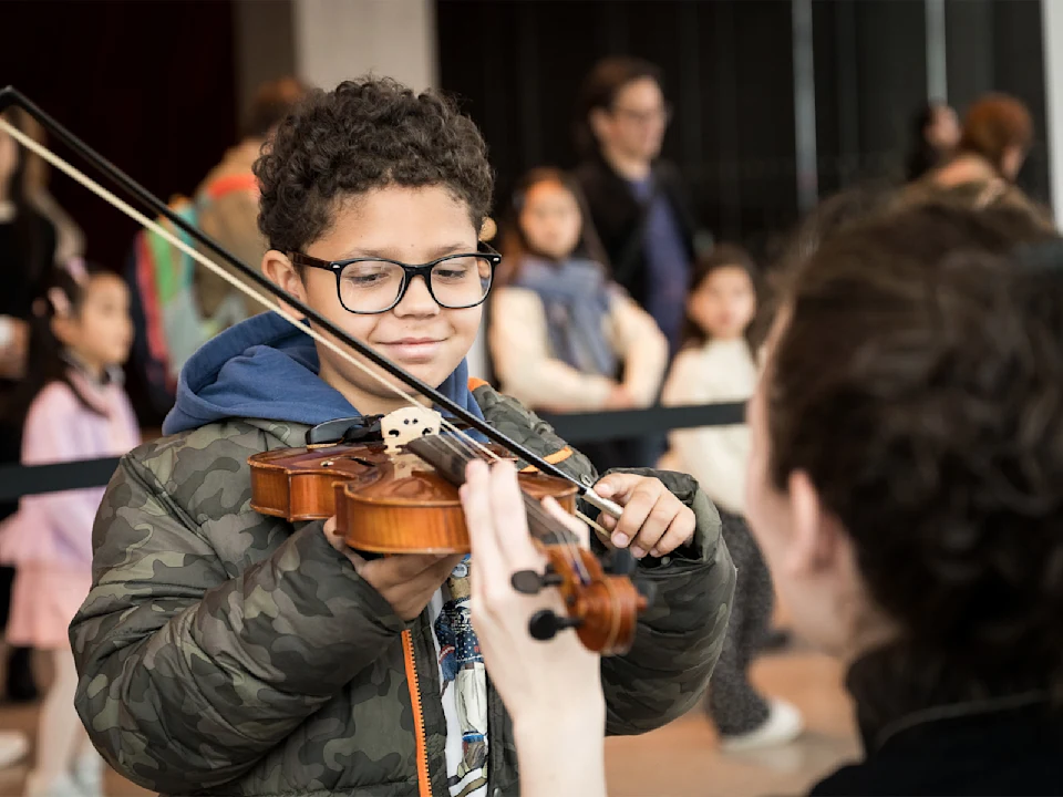 A young boy in glasses plays a violin with help from an adult, while other people watch in the background.