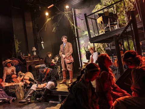 A theatrical scene with a group of actors on stage, some seated and some standing, surrounded by props and set pieces resembling a forest and multi-level platform.