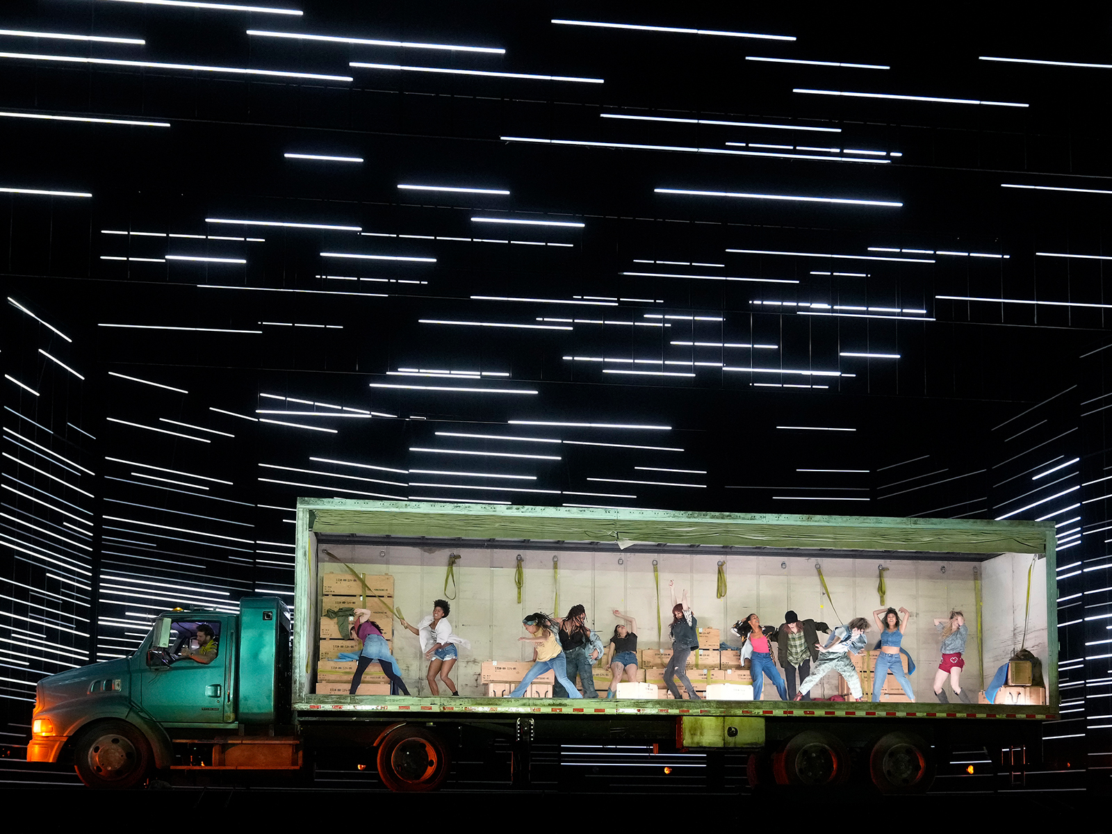 A group of people dance inside the illuminated trailer of a green truck on a stage with horizontal white light lines in the background.