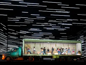 A group of people dance inside the illuminated trailer of a green truck on a stage with horizontal white light lines in the background.