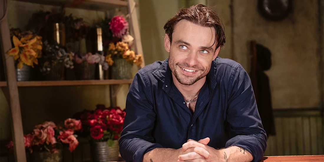 A man with brown hair and a beard, wearing a dark blue shirt, sits at a table and smiles. Behind him are shelves with various colorful flower arrangements.