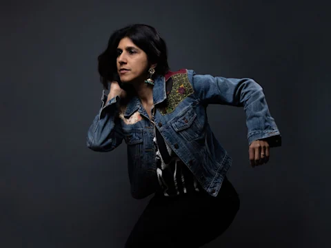 A person with long dark hair poses in a decorated denim jacket against a dark background, with one arm bent and the other extended.