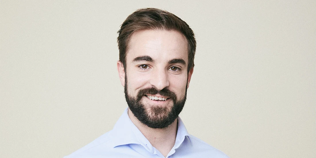 A man with brown hair and a beard wearing a light blue collared shirt, smiling and facing the camera against a plain background.