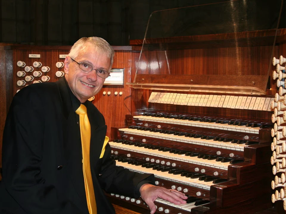 Man in a suit with a yellow tie sits at a large organ console, smiling at the camera.