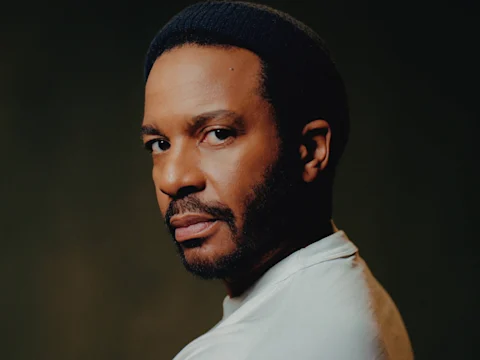 A man with a short beard and mustache wearing a dark knit cap and a light-colored shirt looks over his shoulder against a dark background.