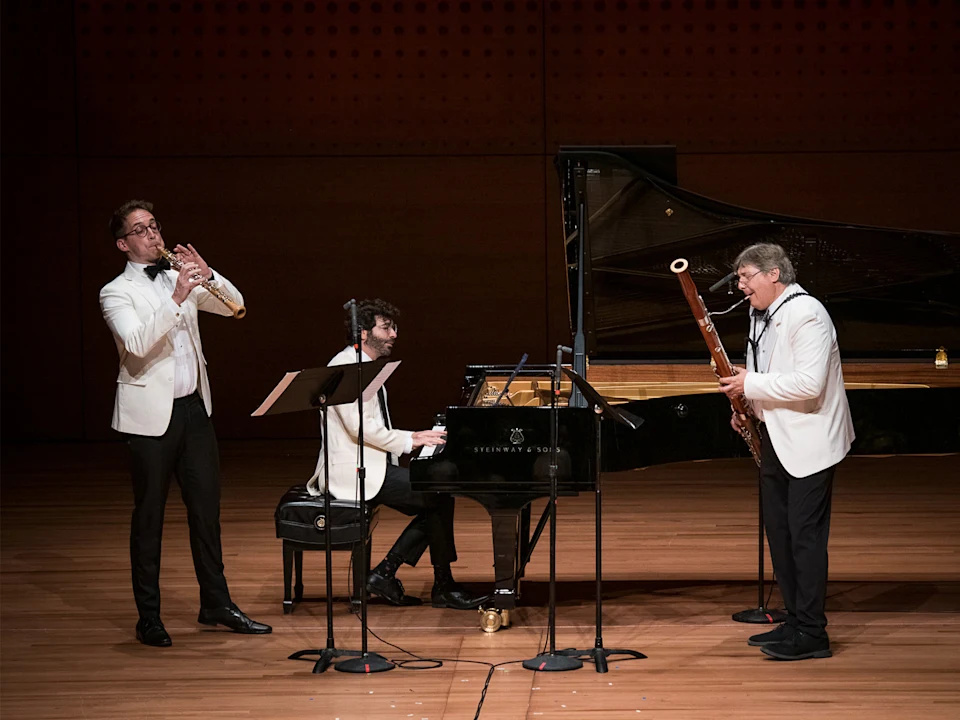Three musicians in white jackets perform on stage with a piano, clarinet, and bassoon. Music stands and microphones are present. The setting appears to be a concert hall.