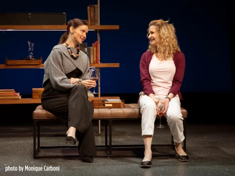 Two women sitting and conversing in a room with shelves and books in the background. Both are holding wine glasses. Photo by Monique Carboni.