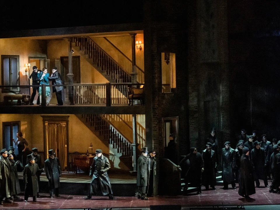 A multi-level stage set shows actors in period costumes performing a scene in a house with stairs and multiple rooms, while others dressed in dark coats and hats gather on the ground floor and outside.