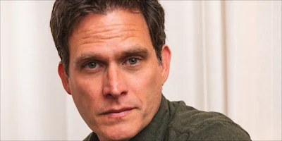 A man with short brown hair and a green shirt looks slightly to the side in front of a beige curtain background.