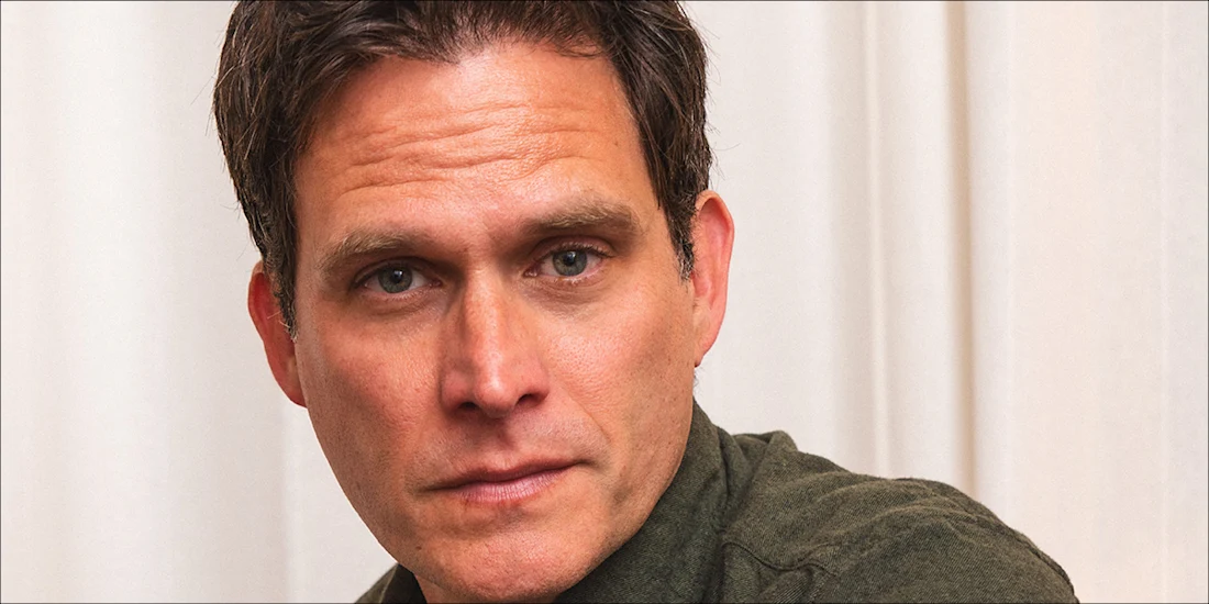 A man with short brown hair and a green shirt looks slightly to the side in front of a beige curtain background.