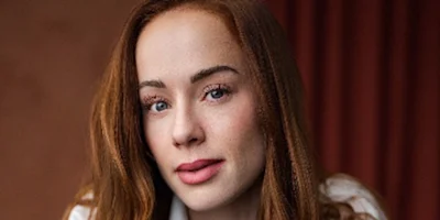 A person with long red hair and fair skin looks at the camera with a neutral expression. The background is out of focus and features warm brown tones.