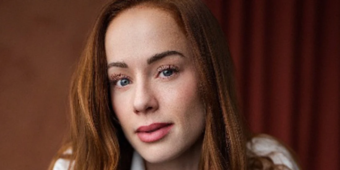 A person with long red hair and fair skin looks at the camera with a neutral expression. The background is out of focus and features warm brown tones.