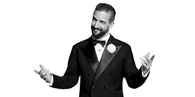 A man in a tuxedo with a bow tie and boutonniere stands with arms outstretched, smiling, against a white background.