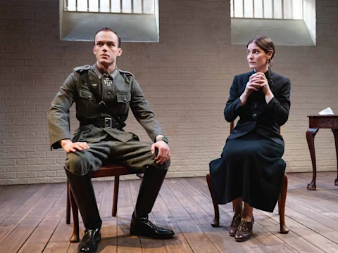 Two actors are seated on a stage, one in a military uniform and the other in a black dress, against a brick wall with barred windows.