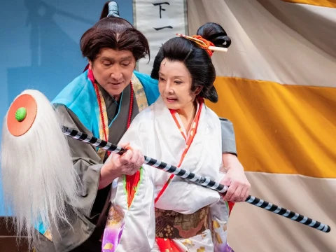 Two actors in traditional Japanese costumes perform on stage. The man assists the woman in holding a long staff.