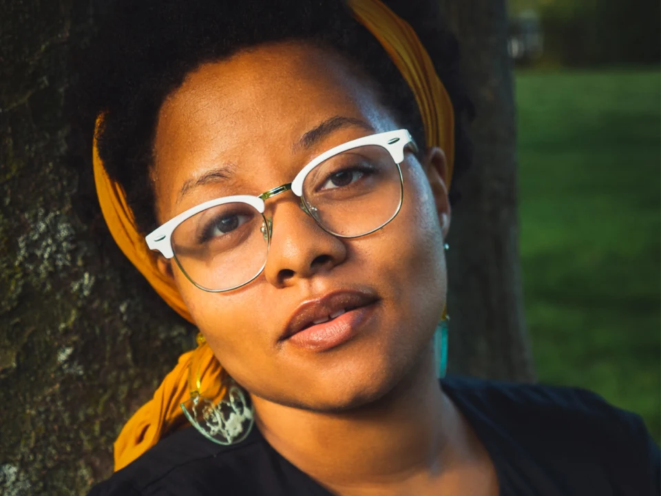 Person with glasses and a yellow headband, wearing a black shirt, leaning against a tree outdoors.