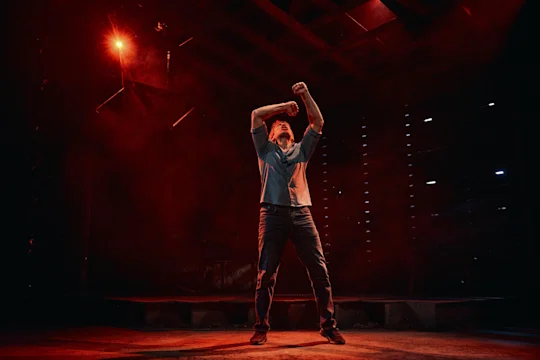 A person stands on a dimly lit stage with red lighting, raising both arms and tilting their head back, surrounded by a dramatic atmosphere.