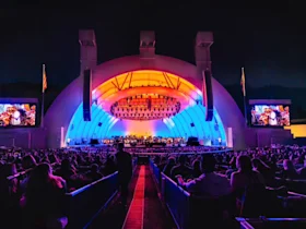 Photo of Hollywood Bowl amphitheater with a large crowd watching a live performance on an illuminated outdoor stage with arched architecture and large screens at night.