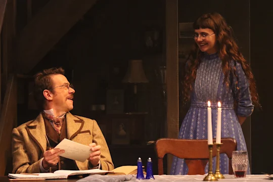A man in a suit sits at a table holding papers, looking at a woman in a blue dress standing and smiling beside him; two lit candles are on the table.