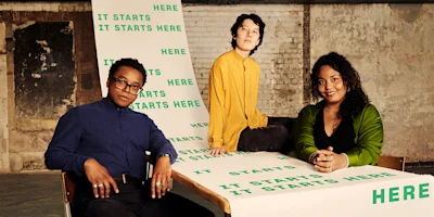 Three people pose indoors with a large banner that reads "IT STARTS HERE" multiple times, set against a worn brick wall background.