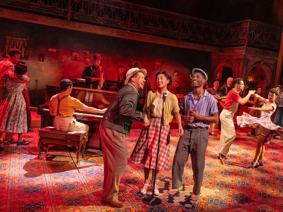 Performers sing and dance on a colorful stage set in a lively, vintage-themed musical scene with a pianist and an energetic crowd.
