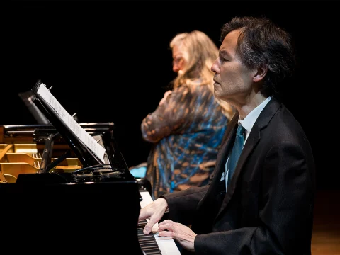 A man in a suit plays the piano while a woman with long hair, seated nearby, looks intently at music sheets. Both are on a dimly lit stage.