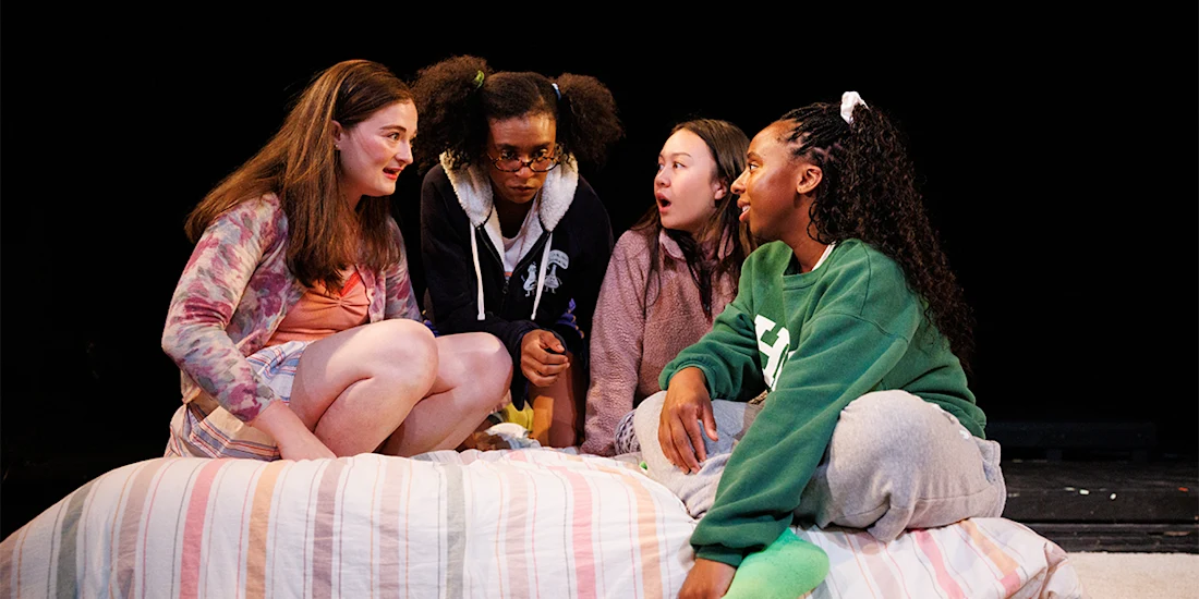 Four teenage girls sit closely together on a bed, engaged in conversation, with a dark background behind them.