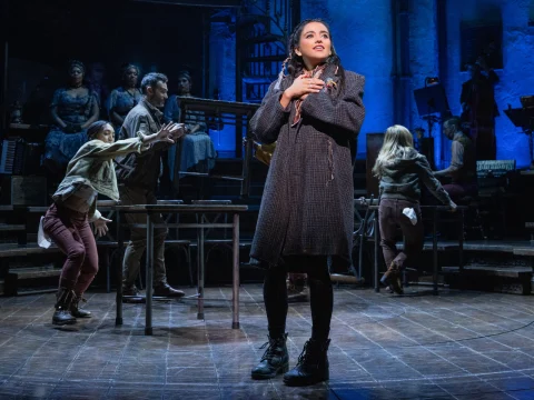 A woman stands center stage with hands near her chest, surrounded by other performers in an intricate theatrical set, with blue lighting highlighting the scene.