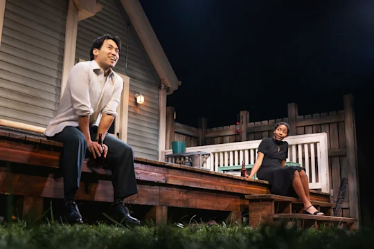 A man sits on the edge of a wooden porch smiling, while a woman in a black dress sits farther back on a bench, looking at him. A house and wooden fence are in the background.