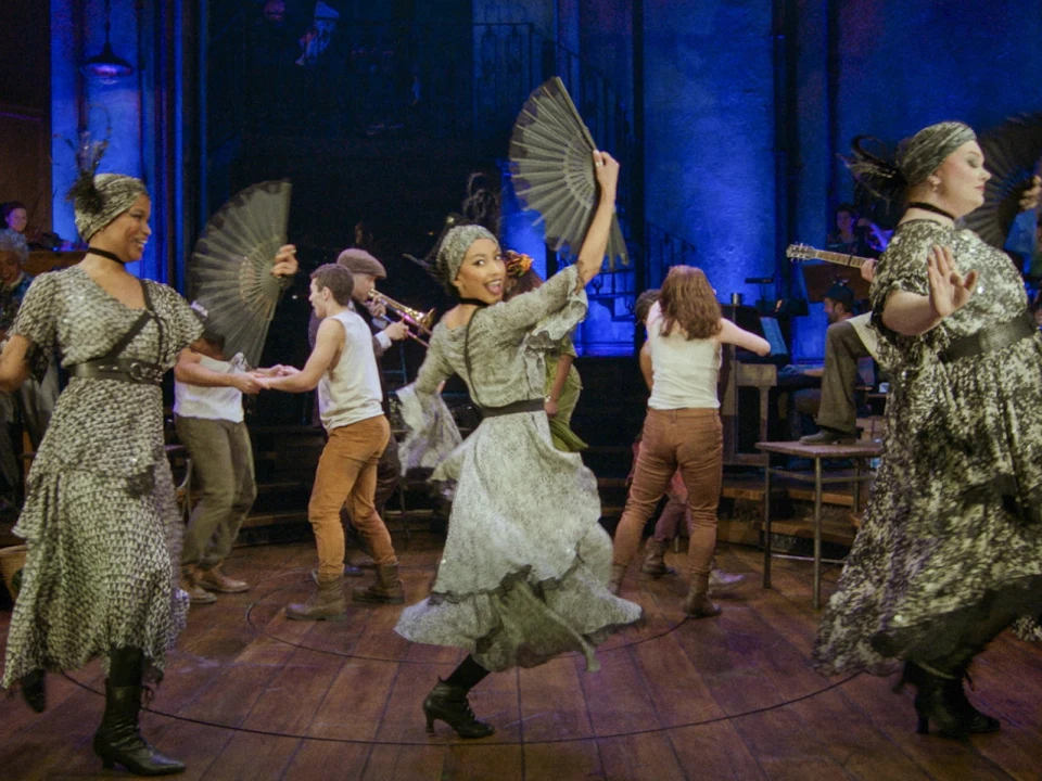 A group of people in historical costumes dance and play instruments on a wooden stage, illuminated by blue lighting.