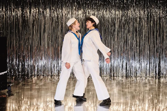 Two people in sailor costumes stand face-to-face and link arms on a shiny stage with a silver tinsel backdrop.