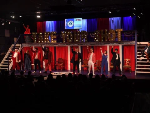 A stage performance with actors in period costumes stands, each raising a hand. A sign above them reads "Take Your Shot" with illuminated letters. The audience is visible in the foreground.