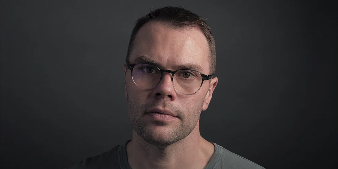 A man with short hair and glasses wearing a grey shirt looks at the camera against a dark, plain background.