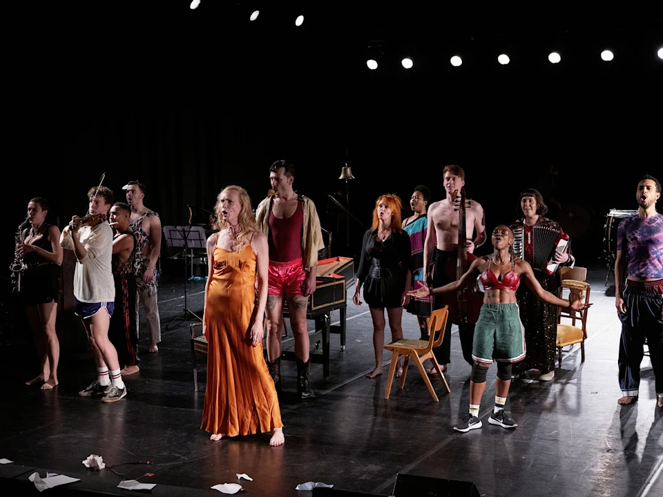 A diverse group of performers in varied costumes stand on a stage under bright lights, facing the audience. Musical instruments and scattered chairs are visible.