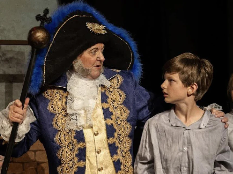 An older person in elaborate historical attire with a hat and staff appears to be speaking to a young boy in a simple shirt.