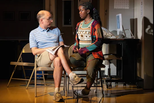 A man and a young woman sit on chairs facing each other in a room, having a serious conversation. The man holds papers, and a computer is visible in the background.